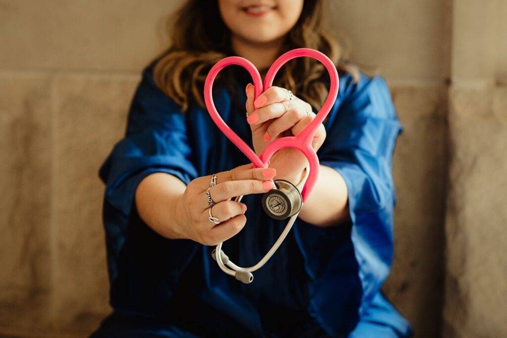 a woman in dark blue scrubs holding a red stethoscope towards the camera and forming a heart shape with the lines of the scope